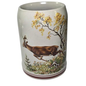 May include: A ceramic mug with a cream-colored background and a brown and yellow deer and tree design. The mug has a red stripe around the base and is cylindrical in shape.