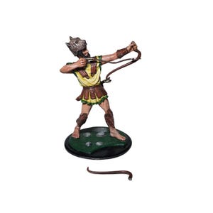 May include: A figurine of a warrior in a yellow and brown tunic, aiming a bow. The warrior wears a helmet with a wolf's head and stands on a green and black base. The figurine is a collectible item.