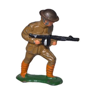 May include: A vintage toy soldier figurine in a tan uniform, brown helmet, and orange boots, holding a black machine gun. The figurine stands on a green base, with visible paint wear, suggesting its age and use.