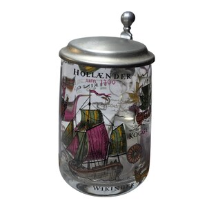 May include: A clear glass beer stein with a metal lid and a thumb lift. The exterior features colorful illustrations of historical sailing ships, including a ship labeled KOGGE, with text reading HOLLANDER um 1700 and WIKINGER printed on the glass.