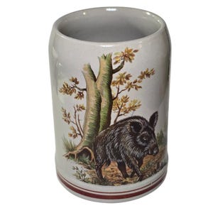 May include: A ceramic mug with a forest scene featuring a brown wild boar, a tree, and autumn foliage. The mug has a cream-colored background with a red stripe near the base.