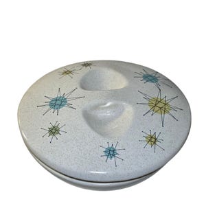May include: A round ceramic casserole dish lid with a speckled white finish. The surface features a pattern of black starburst lines with light blue and yellow circular accents. The lid has two recessed, teardrop-shaped indentations for gripping.