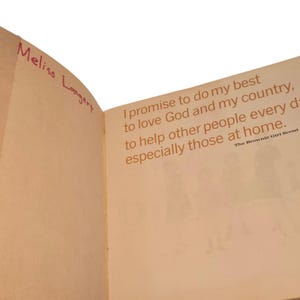 May include: Open book pages with text in orange. The text reads "I promise to do my best to love God and my country, to help other people every day, especially those at home." The name "Meliss Langert" is written in pink.
