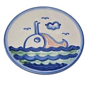 May include: A round ceramic plate with a hand-painted design of a whale in the ocean. The whale is blue and tan, with blue water and a blue sky. The plate has a blue border and is on a white background.