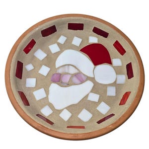May include: A round, mosaic-tiled bowl featuring a Santa Claus design. The bowl has a tan interior with red, white, and pink glass tiles forming Santa's hat, beard, and face. The rim is a warm brown color.
