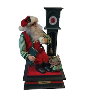 May include: A Santa Claus figurine sitting on a wooden base with a clock tower behind him. He is holding a mug that says "Santa" and a book. The base has the word "Noel" on it.