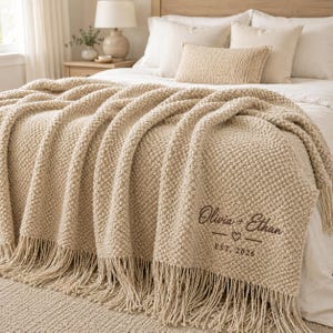 May include: A beige chunky knit throw blanket with fringe trim, featuring dark brown embroidered text that reads Olivia + Ethan with a small heart symbol and the text EST. 2026, draped over a bed with white bedding and decorative pillows.