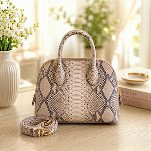 May include: A dome-shaped handbag with a snakeskin pattern in shades of beige, gray, and brown. The bag has a top handle and a detachable shoulder strap. Gold-toned zippers and hardware add a touch of elegance. The bag is sitting on a wooden surface.