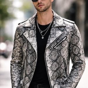 Puede incluir: Chaqueta de cuero con estampado de piel de serpiente en tonos grises y blancos, con un estilo motero clásico con cuello de solapa, cremallera asimétrica y múltiples bolsillos. Se lleva con una camiseta negra y gafas de sol.