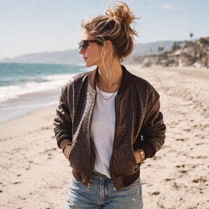 May include: A brown bomber jacket with a textured pattern, worn over a white t-shirt and blue jeans. The jacket has a ribbed collar and cuffs. The person is wearing sunglasses and jewelry, standing on a sandy beach with the ocean in the background.