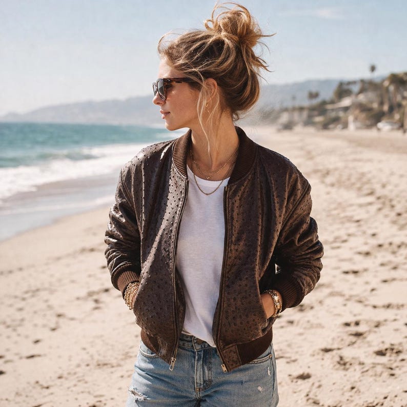 May include: A brown bomber jacket with a textured pattern, worn over a white t-shirt and blue jeans. The jacket has a ribbed collar and cuffs. The person is wearing sunglasses and jewelry, standing on a sandy beach with the ocean in the background.