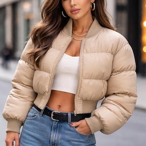 Puede incluir: Chaqueta acolchada beige con cierre de cremallera y puños acanalados. La chaqueta se combina con un top corto blanco, vaqueros azules y un cinturón negro. Joyas doradas y un bolso de mano completan el look.