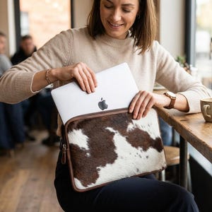 May include: A brown and white cowhide laptop sleeve with a zipper closure. The sleeve is designed to hold a laptop, and is being held by a person. The sleeve has a brown leather trim.