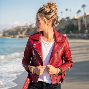 May include: A red snakeskin-textured leather jacket with a zipper closure and silver hardware. The jacket is worn over a white t-shirt. The person is wearing gold bracelets and necklaces. The background shows a beach and ocean.