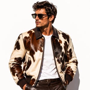 May include: A cowhide bomber jacket with a brown and cream pattern. The jacket has a black collar and cuffs, and a zipper closure. The model is wearing sunglasses and a white t-shirt.