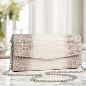 May include: A light grey and white clutch bag with a silver chain strap. The bag has a textured, crocodile-like pattern. The chain is draped across a white surface. The background is blurred, with a vase of flowers visible.