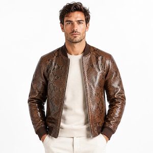 May include: Brown snakeskin-patterned leather bomber jacket with a ribbed collar, cuffs, and hem. The jacket has a zipper closure and a classic bomber silhouette. The model is wearing a cream-colored sweater and white pants.
