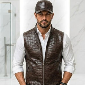 May include: A brown leather vest with a crocodile skin pattern, paired with a matching baseball cap. The vest has a zipper closure and a white collared shirt underneath. A black wristwatch is visible.