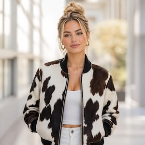 May include: A cowhide print bomber jacket with a black collar and cuffs. The jacket features a white and brown pattern and a front zip. The model is wearing a white top and gold jewellery.
