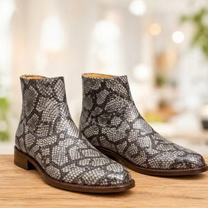 May include: A pair of ankle boots with a snakeskin pattern in shades of gray and black. The boots have a low heel and a rounded toe. The boots are made of leather and have a zipper closure. The boots are displayed on a wooden surface.
