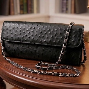 May include: A black clutch with a silver chain strap. The clutch has a textured surface with a pattern of small bumps. The bag is rectangular with a flap closure. The chain strap is draped across a wooden table.