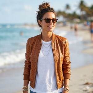 May include: A brown leather bomber jacket with a textured pattern. The jacket has a ribbed collar and cuffs. The person is wearing a white t-shirt and sunglasses. The background is a beach with the ocean and palm trees.