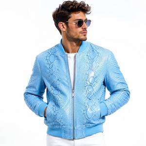 Könnte beinhalten: Hellblaue Bomberjacke mit Schlangenmuster. Die Jacke hat einen Reißverschluss, gerippte Bündchen und einen gerippten Saum. Die Jacke wird mit einem weißen T-Shirt und einer weißen Hose getragen. Das Model trägt eine Sonnenbrille.