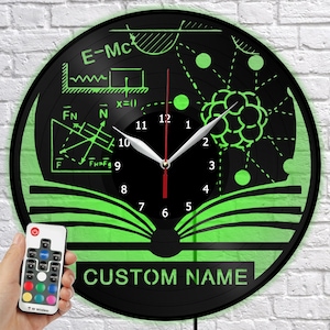 Puede incluir: Reloj de pared negro con diagramas científicos verdes y el texto "CUSTOM NAME". La esfera del reloj presenta ecuaciones, un modelo atómico y un diseño de libro. Incluye un mando a distancia.