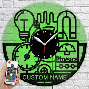May include: A circular wall clock made from a vinyl record featuring black cut-out science icons including a lightbulb, magnet, and gauge. The center has a black clock face with white numbers and hands. The text Physics and CUSTOM NAME are visible.