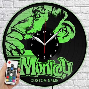 Puede incluir: Reloj de vinilo negro con un diseño de mono verde y la palabra "Monkey". El reloj tiene manecillas blancas y un mando a distancia con opciones de color. El diámetro del reloj es de aproximadamente 30 cm.