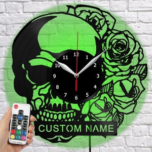 Könnte beinhalten: Schwarze, schädelartige Wanduhr mit grünen Akzenten und einem floralen Design. Das Zifferblatt ist schwarz mit weißen Zeigern und Zahlen. Die Worte "CUSTOM NAME" stehen unten. Eine Fernbedienung befindet sich unten links.