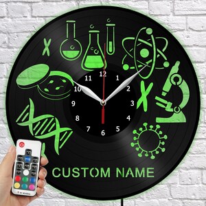 Puede incluir: Un reloj de vinilo negro con un tema científico verde brillante. El reloj presenta equipos de laboratorio, ADN y un microscopio. El texto "CUSTOM NAME" está en la parte inferior. Un mando a distancia está en la esquina inferior izquierda.