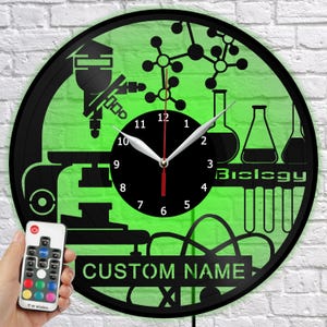 Könnte beinhalten: Eine schwarz-grüne Wanduhr zum Thema Biologie mit einem Mikroskop, Molekularstrukturen und Laborgeräten. Das Zifferblatt hat einen schwarzen Hintergrund mit weißen Zahlen und Zeigern. Der Text "Biology" und "Custom Name" sind sichtbar.