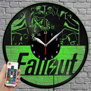 May include: A circular wall clock crafted from a black vinyl record featuring a green illuminated graphic of a power armor helmet from the Fallout video game series. The clock face displays the word Fallout in large black letters with a lightning bolt.