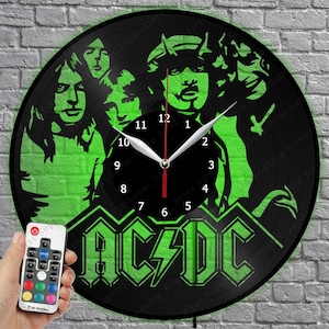 May include: A circular wall clock crafted from a black vinyl record featuring a green backlit silhouette of the band AC/DC. The clock face displays white numbers from 1 to 12 with white hour and minute hands and a red second hand. Includes a remote.