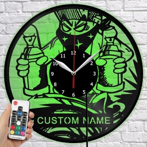 May include: A black and green wall clock with a graffiti-style design. The clock features a cartoon character holding spray paint cans, with the text "CUSTOM NAME" at the bottom. The clock is round and has a black clock face with white hands. A remote control is in the image.