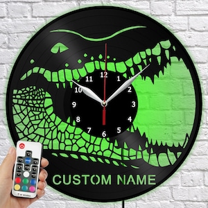 Puede incluir: Un reloj de vinilo negro con un diseño de caimán verde. La esfera del reloj tiene manecillas blancas y un segundero rojo. El texto "CUSTOM NAME" está en la parte inferior. Un mando a distancia está en la esquina inferior izquierda.
