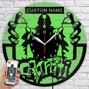 May include: A black and green wall clock with a graffiti artist design. The clock features a black clock face with white numbers and hands, and the text "CUSTOM NAME" above the clock. A remote control is in the lower left corner.