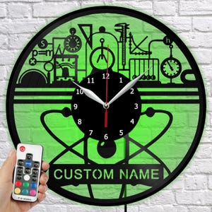 Puede incluir: Un reloj de pared negro y verde con temática científica. El reloj tiene una esfera negra con manecillas blancas y un segundero rojo. El reloj tiene un fondo verde con gráficos relacionados con la ciencia y el texto "CUSTOM NAME". Se ve un mando a distancia.