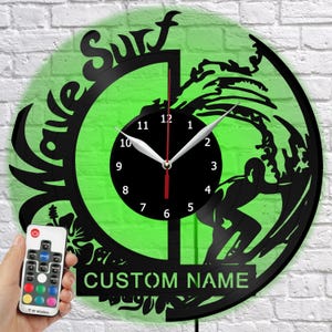 May include: A circular wall clock crafted from a black vinyl record featuring a cutout silhouette of a surfer riding a wave, the text "WAVE SURF" and "CUSTOM NAME," and a central black clock face with white numbers and a red second hand.