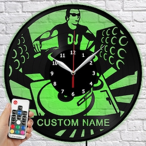 Op de afbeelding: Zwarte vinylplatenklok met een groen neon DJ-ontwerp. De klok heeft een silhouet van een DJ, draaitafels en de tekst "CUSTOM NAME". Een afstandsbediening met kleuropties wordt in de linkerbenedenhoek vastgehouden.
