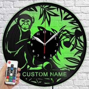 Puede incluir: Un reloj de pared negro y verde con temática de mono con opción de nombre personalizado. El reloj presenta una silueta de mono, follaje y una esfera negra con manecillas blancas. Incluye un mando a distancia.