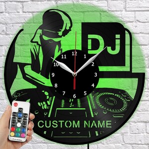 Puede incluir: Reloj de pared con temática de DJ en negro y verde. El reloj está hecho de un disco de vinilo y presenta una silueta de DJ, la palabra "DJ" y el texto "CUSTOM NAME". Se sostiene un mando a distancia con opciones de color en la mano.