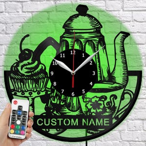 May include: A black wall clock with a green background, featuring a cupcake, teapot, and teacup design. The clock has a black face with white hands and the text "CUSTOM NAME" at the bottom. A remote control is held in the hand.