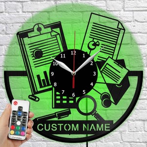 Può includere: Un orologio da parete circolare con un design a silhouette nera di articoli da ufficio, tra cui una cartellina, documenti con grafici, una lente d'ingrandimento e una penna. Il quadrante è nero con numeri bianchi e include il testo CUSTOM NAME.