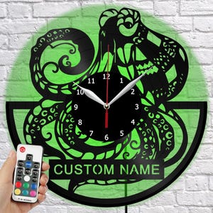 May include: Black octopus-themed wall clock with a green illuminated background. The clock features a black face with white hands and numbers. The text "CUSTOM NAME" is displayed on the bottom. A remote control is held in the hand.