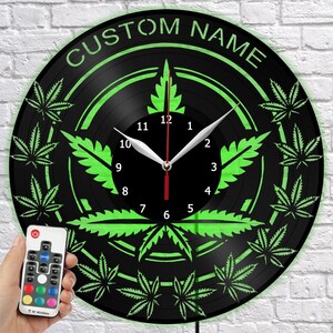 May include: A black vinyl record clock with a green cannabis leaf design. The clock face has white hands and a red second hand. The text "CUSTOM NAME" is at the top. A remote control is in the lower left corner.