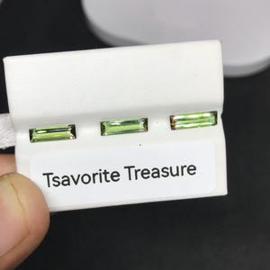 May include: Three rectangular, light green tsavorite gemstones are displayed on a white card with the text "Tsavorite Treasure." The gemstones are set in a row, showcasing their clarity and color. The card is held by a hand.