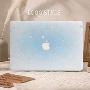 May include: A light blue laptop hard shell case featuring a subtle pattern of small white stars and faint celestial sparkles. The centre of the case has a precise cutout revealing the silver Apple logo. The surface has a smooth, matte finish.