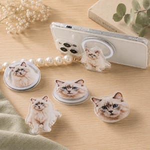Peut inclure: Une collection de supports de téléphone circulaires présentant l'image imprimée d'un chat duveteux de couleur claire aux yeux bleus. Un support est fixé au dos d'une coque de smartphone transparente, et trois autres sont présentés séparément.